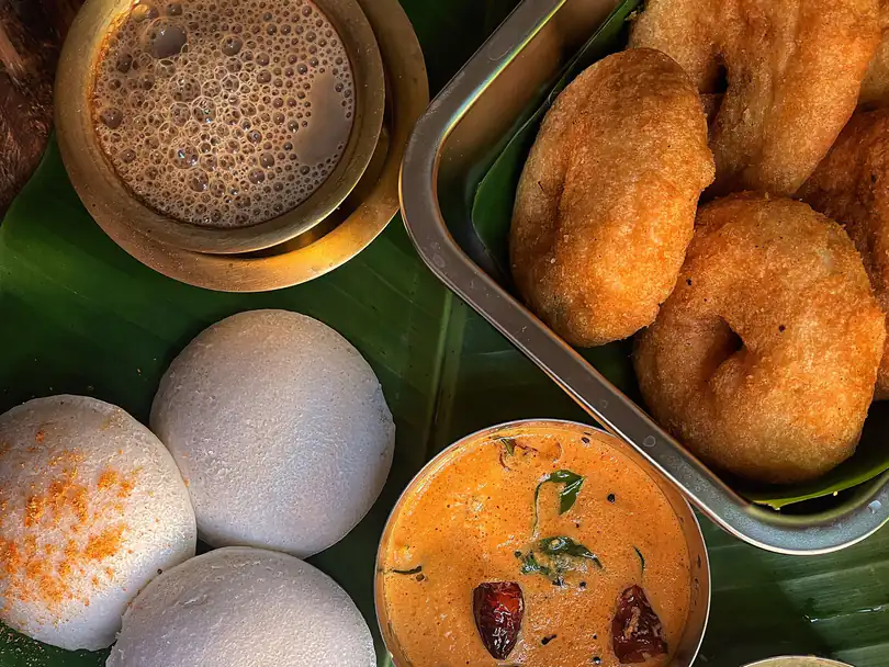 Idli dusted with podi, a tray of medu vadai, tomato chutney, coconut chutney, small bananas and a brass tumbler of filter coffee on a banana leaf.