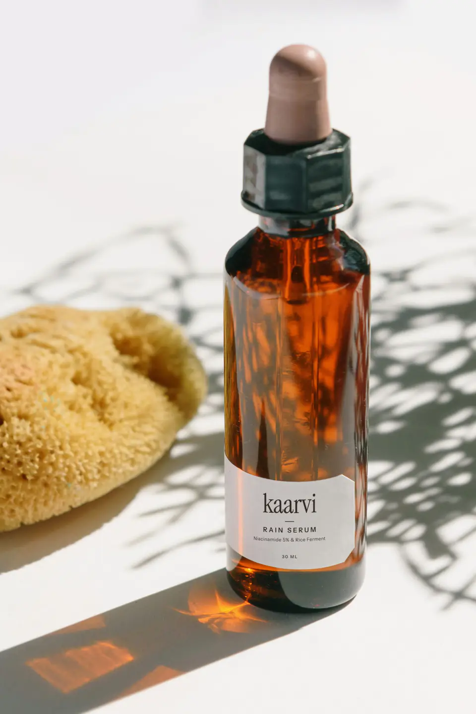 Kaarvi Rain Serum in an amber glass dropper bottle, standing in morning sun beside a natural sponge